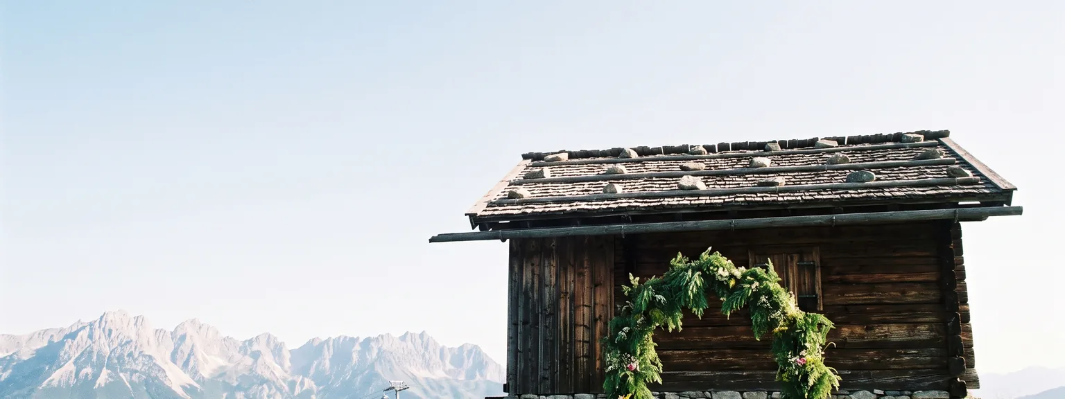 Almhütte aus dunklem Holz mit steinbeschwertem Schindeldach auf einer Bergwiese, davor ein grüner Blätterbogen, im Hintergrund Kalkgipfel und eine Gondel.