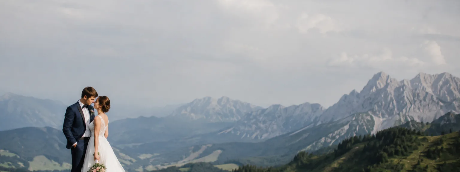 Paar in Hochzeitskleidung auf einer Bergwiese, dahinter eine Alpenkette.